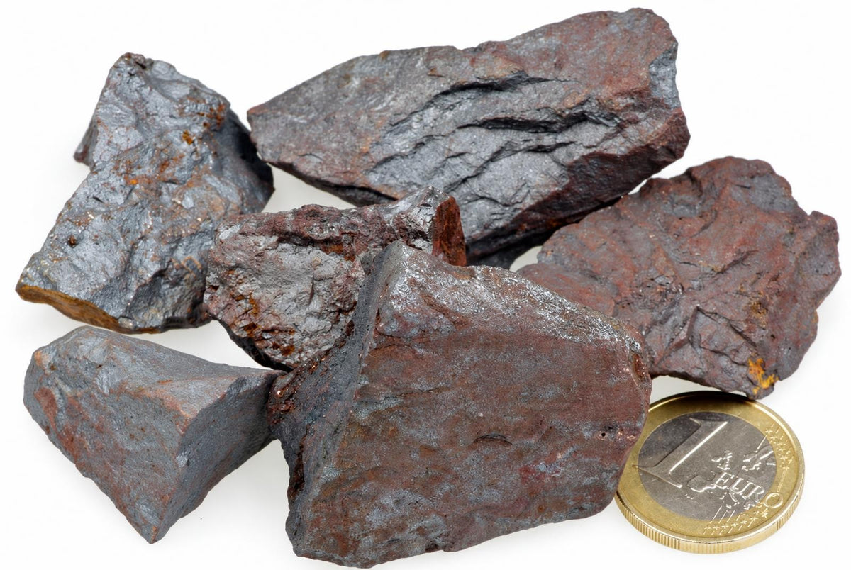 A pile of dark grey and reddish-brown raw hematite rocks on a white surface, next to a one euro coin.