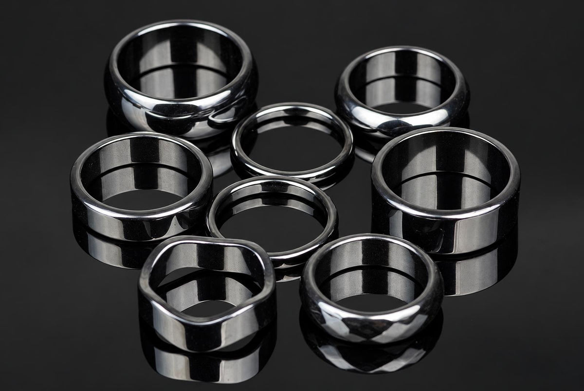 Seven silver-toned rings with varied shapes and faceted surfaces arranged on a dark, reflective surface.