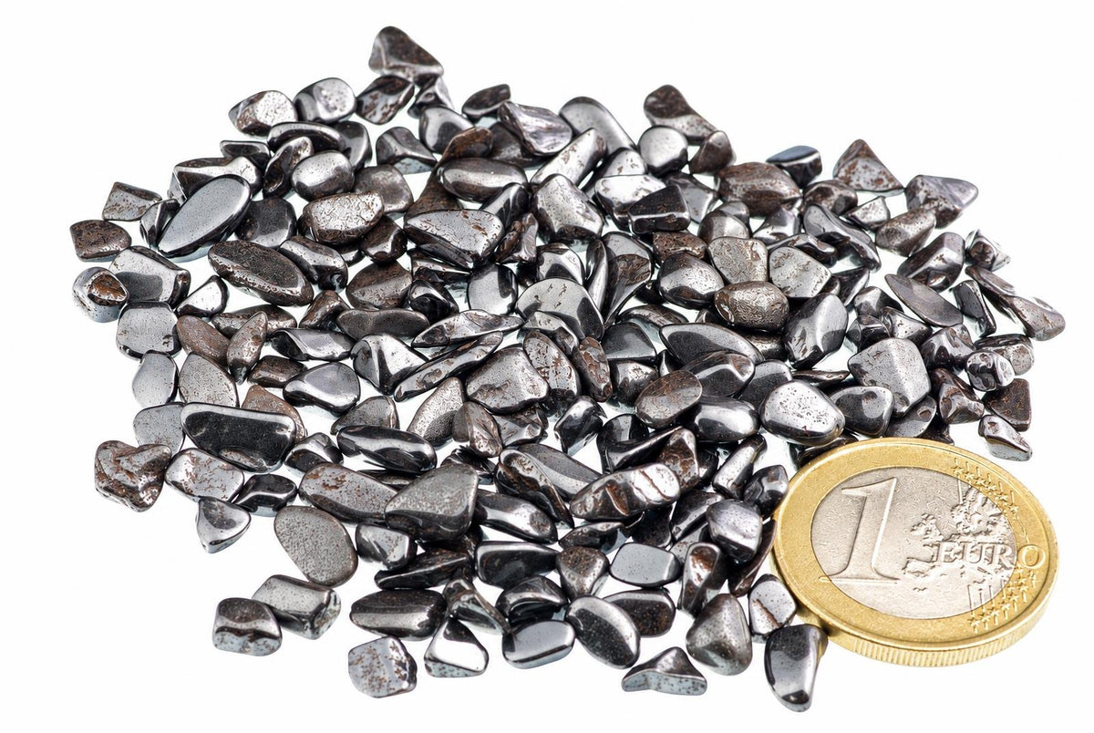 A pile of small, dark grey and silver-toned Hematite stones on a white surface with a one Euro coin for scale.