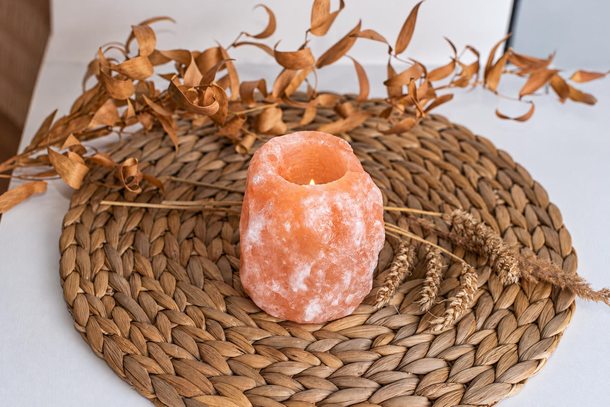 A lit Himalayan rock salt candle sits on a woven brown mat, surrounded by dried leaves and wheat stalks.