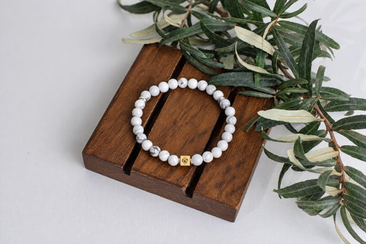 A bracelet with white and grey patterned beads, a gold-coloured square charm, and silver-toned spacers rests on a dark wooden surface beside green leaves.