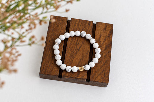 A bracelet with white and grey marbled beads, silver spacers, and a gold-toned square charm rests on a dark wooden stand.
