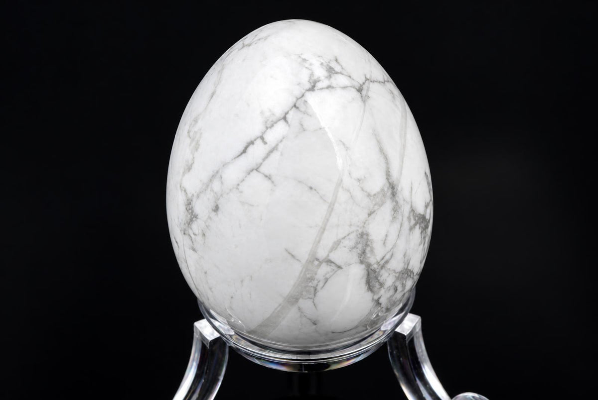 A polished white egg-shaped stone with grey veining sits on a clear stand against a dark background.