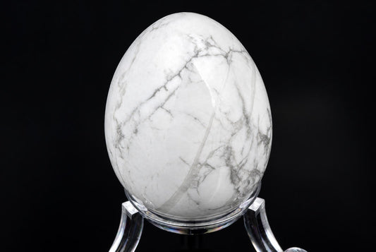 A polished white egg-shaped stone with grey veining sits on a clear stand against a dark background.