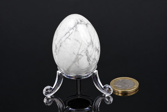 A white egg-shaped stone with grey veins sits on a clear stand next to a coin, against a dark background.