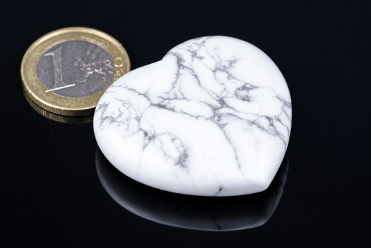 A polished white heart-shaped stone with grey veins sits on a dark surface next to a one-euro coin.