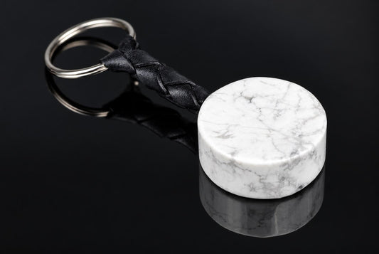 A white cylindrical stone with gray veining is attached to a black braided key strap and metal ring on a reflective black surface.