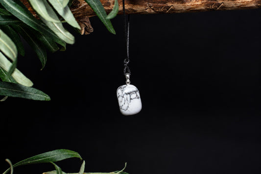 A white pendant with grey veins hangs from a black cord on a dark surface, framed by green leaves.