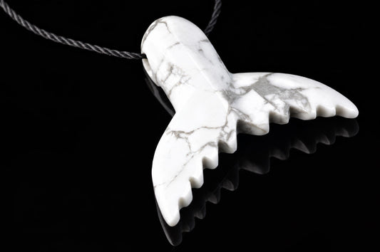 A white pendant with grey veining, carved into a dolphin tail shape, hangs from a dark cord on a black reflective surface.