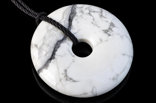 A white pendant with grey veining, shaped like a donut with a central hole, threaded on a dark cord against a black background.