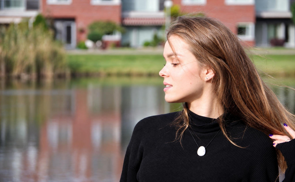 A woman wears a black turtleneck with a white oval pendant on a dark cord, outdoors near water.