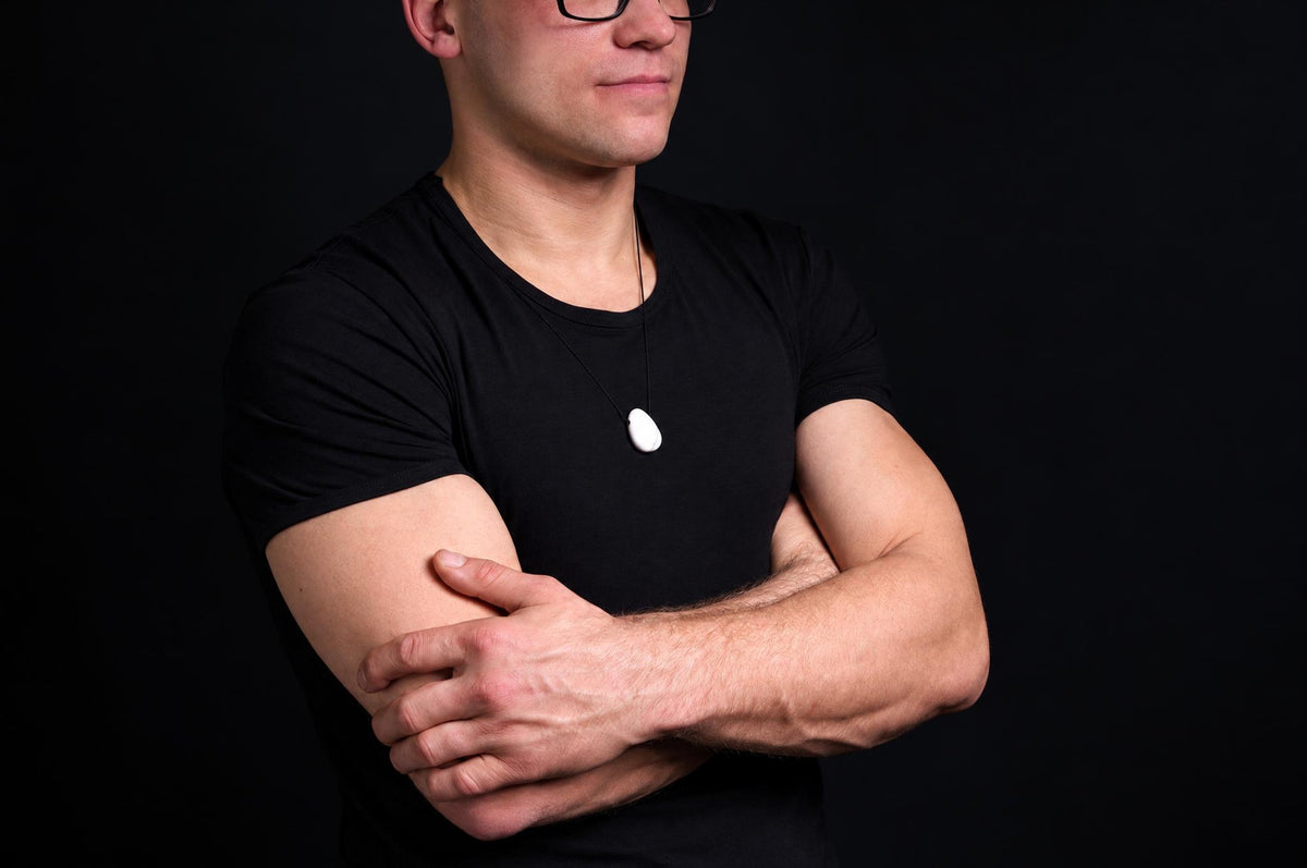 A man in a black t-shirt wears a white pendant on a dark cord against a dark background.