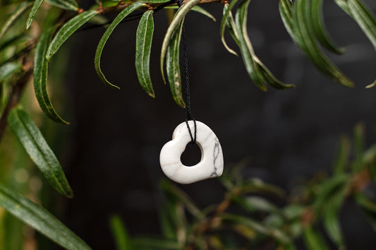 A white pendant with grey veining, shaped like a heart with a hole, hangs from a black cord on a branch with green leaves.