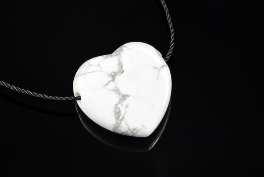 A white heart-shaped pendant with grey veining hangs on a dark cord against a black reflective surface.