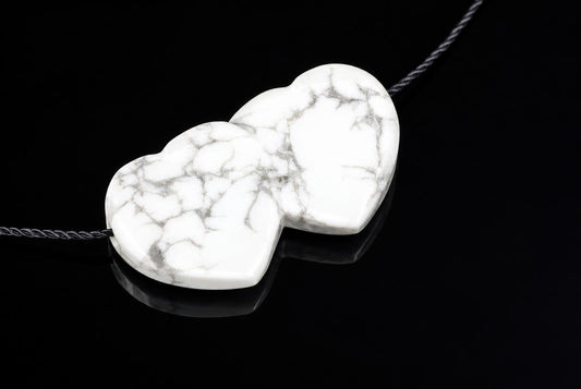 A white pendant with grey veining, shaped like two joined hearts, on a dark cord against a black reflective surface.