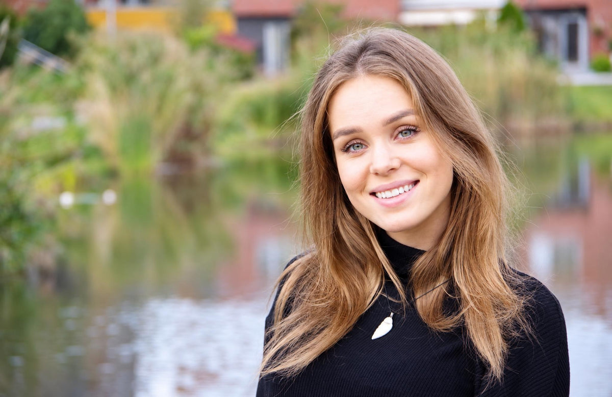A smiling woman with long brown hair wears a black top and a white pendant necklace against a blurred outdoor background.