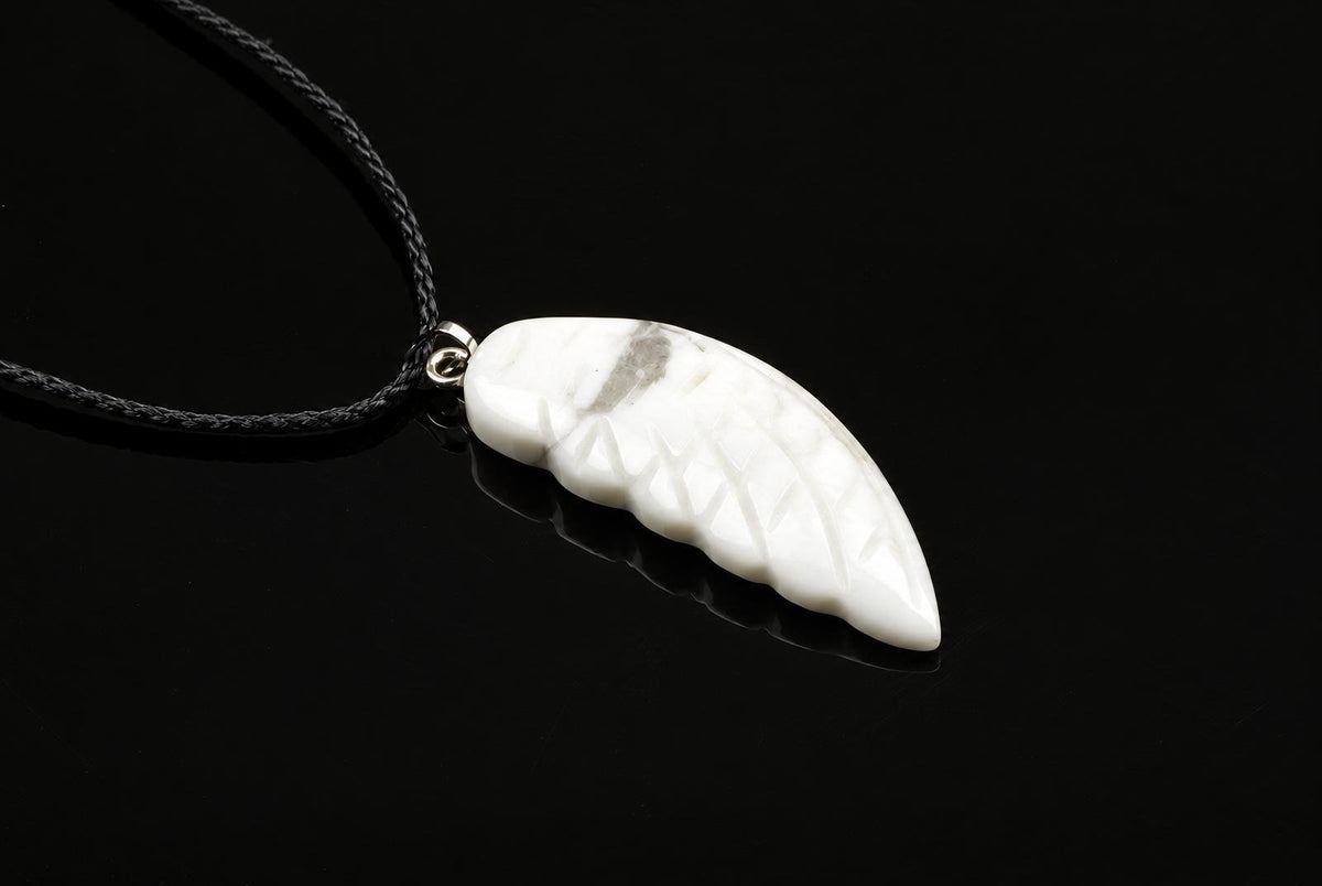 A white pendant shaped like a wing with carved lines and grey veining, attached to a black cord on a dark surface.