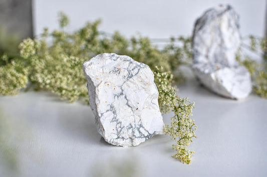 A white, grey-veined Howlite stone stands on a pale surface with small white flowers nearby; another stone is blurred in the background.