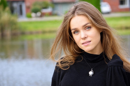 A woman with long brown hair wears a black turtleneck and a silver-toned sphere pendant necklace against a blurred outdoor background.