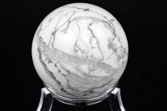 A polished white and grey marbled Howlite sphere rests on a clear stand against a black background.