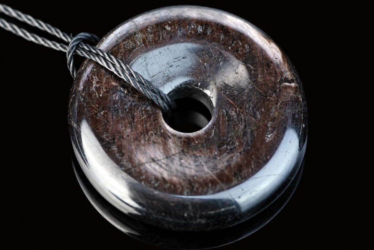 A polished, dark brown donut-shaped pendant with a central hole and silver-toned cord against a black background.