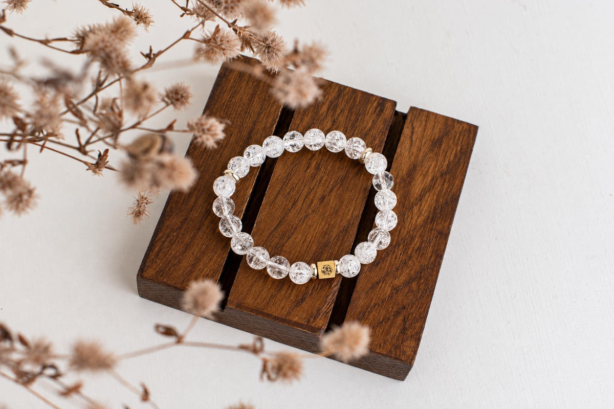 A bracelet with clear, crackled beads and a gold-toned square charm rests on a dark wooden surface.
