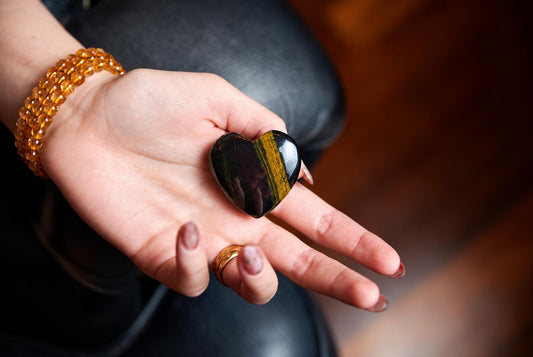 A hand holds a polished heart-shaped stone with dark and yellow stripes, resting on a dark surface.