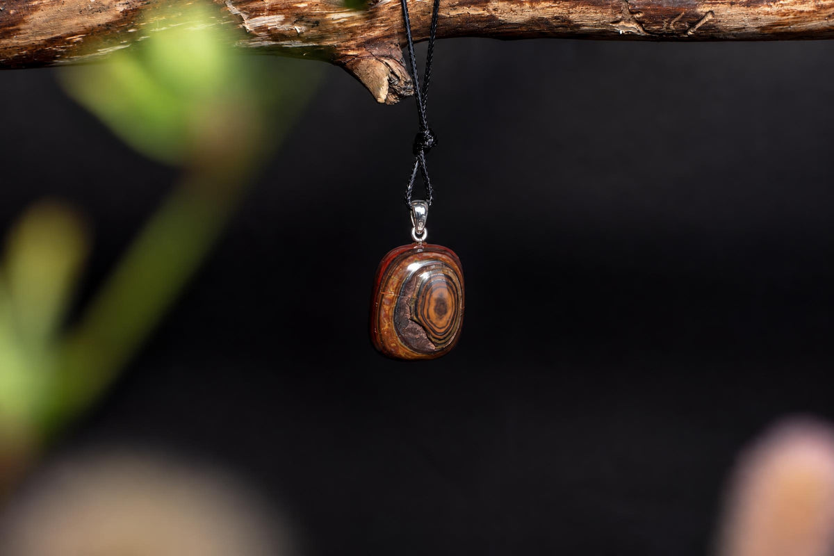 A polished, oval pendant with concentric brown and orange bands hangs from a black cord on a wooden branch against a dark background.