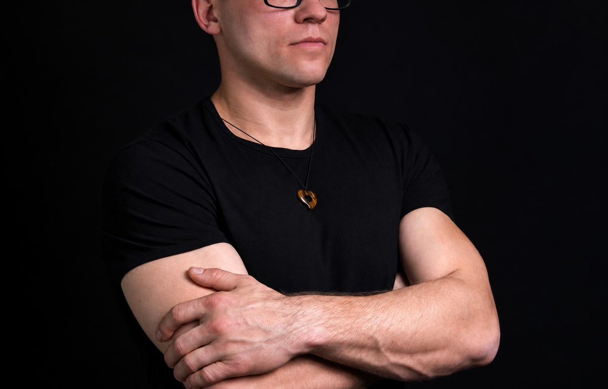 A person wearing a black t-shirt and glasses, with arms crossed, displaying a brown heart-shaped pendant on a dark surface.
