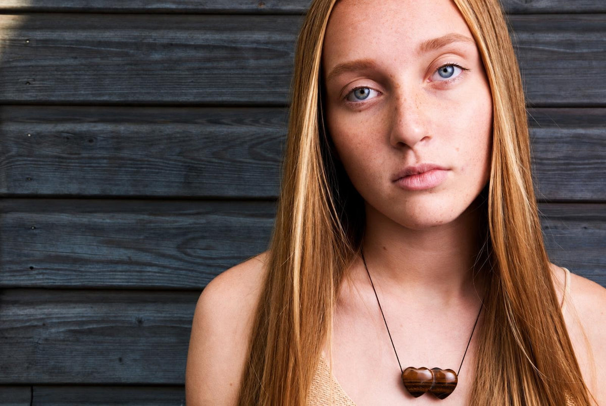 A woman wears a necklace with two dark brown, heart-shaped pendants on a thin cord against a dark wooden background.