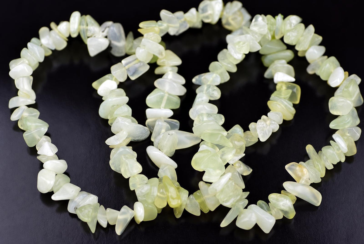 A jade chip bracelet with pale green and white stones arranged in a loose coil on a dark surface.