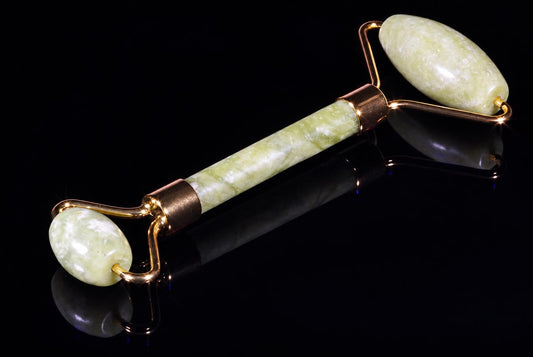 A facial massager roller with a green stone handle and two oval rollers on gold-toned metal frames, resting on a dark reflective surface.