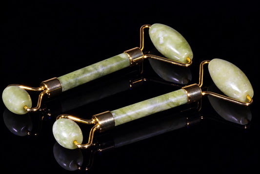 Two jade facial massager rollers with gold-toned metal frames and green stone heads on a dark reflective surface.
