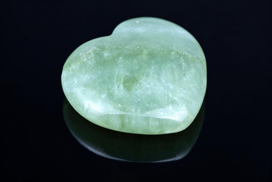 A polished, heart-shaped green stone rests on a dark, reflective surface.