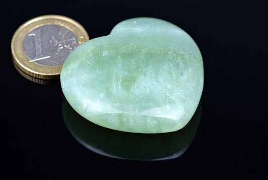 A polished, light green heart-shaped stone rests on a dark surface next to a one-euro coin for scale.