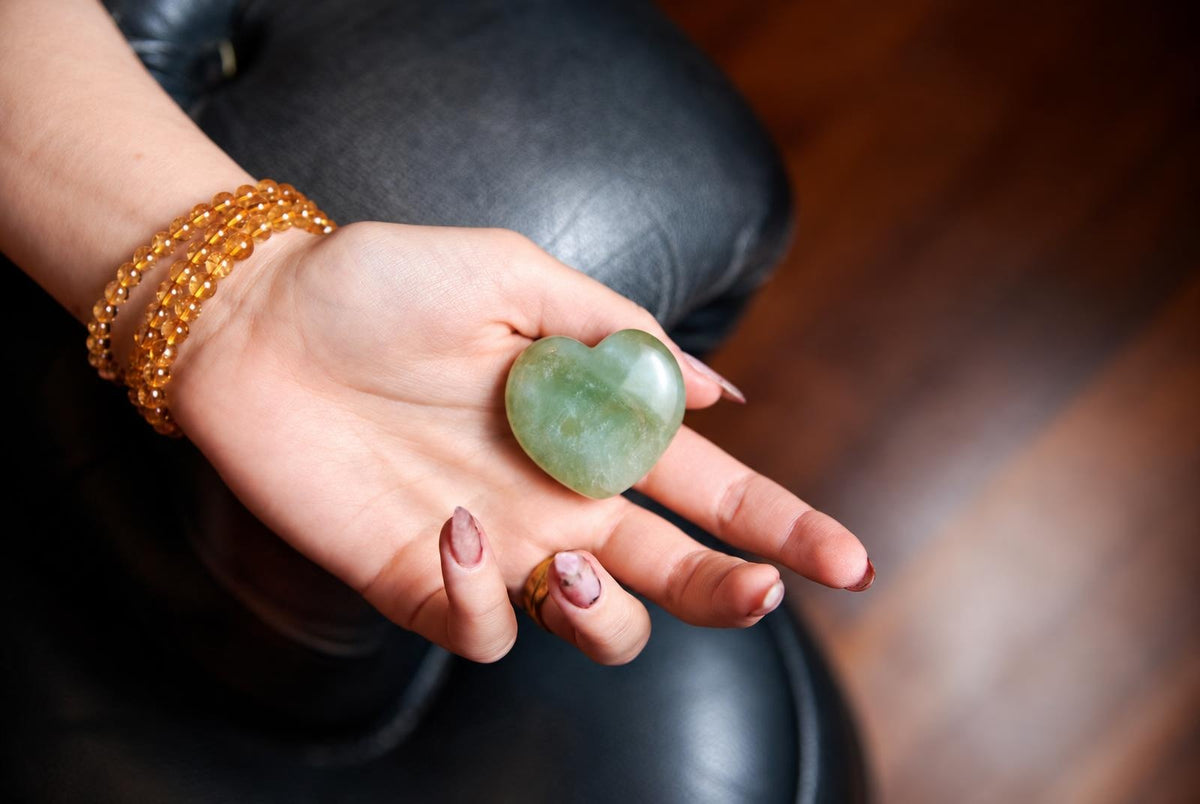 A hand holds a smooth, green heart-shaped stone on a dark surface.