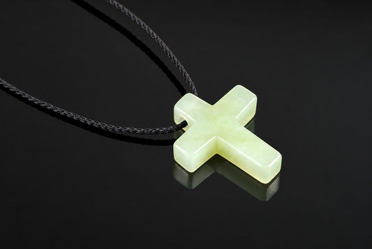 A pale green cross-shaped pendant on a dark cord, resting on a reflective black surface.