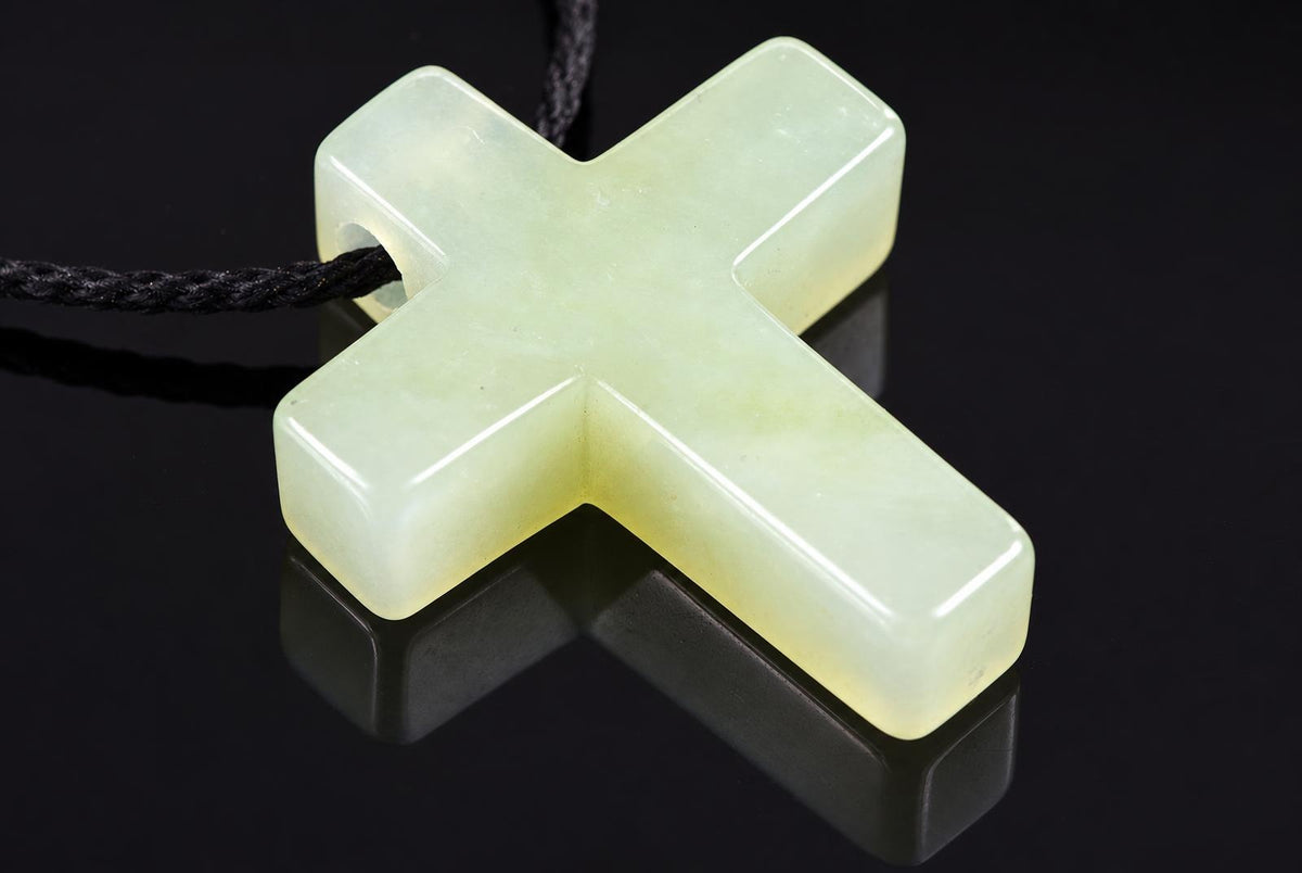 A pale green jade cross pendant with a black cord, resting on a dark reflective surface.