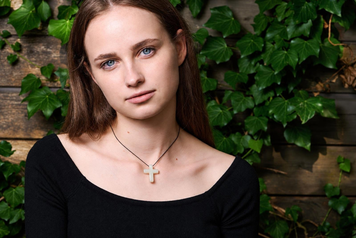 A woman wears a black top and a cross pendant on a dark cord, set against a wooden wall with green ivy.
