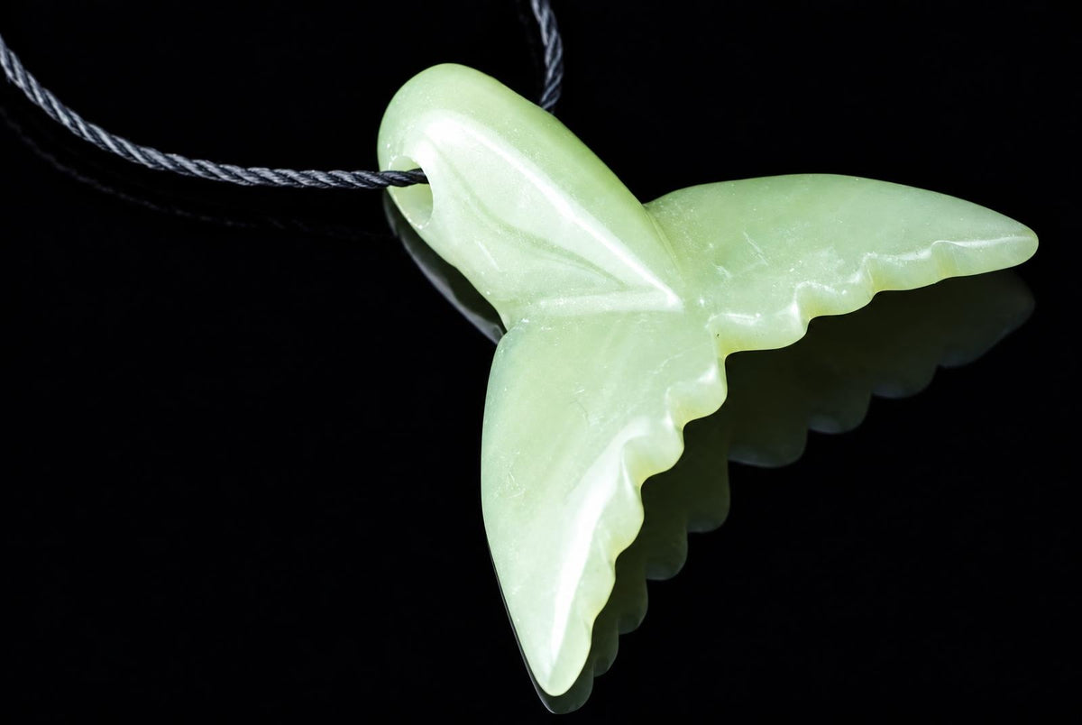 A light green jade pendant shaped like a dolphin tail hangs from a dark cord against a black reflective surface.