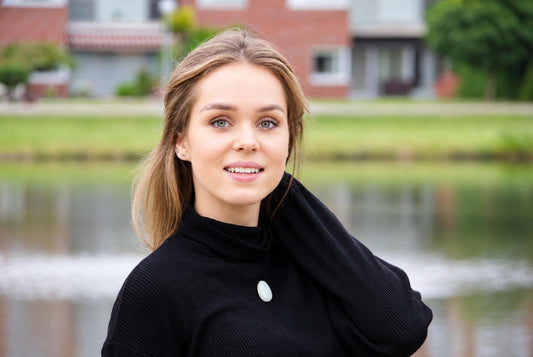 A woman wears a black turtleneck with a pale green oval pendant necklace, standing outdoors near water.