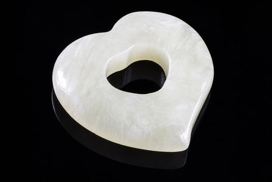 A polished, pale greenish-white heart-shaped pendant with a central hole, resting on a dark surface.
