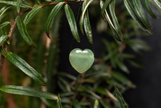 A translucent, pale green heart-shaped pendant hangs from a thin black cord against a dark background with green leaves.