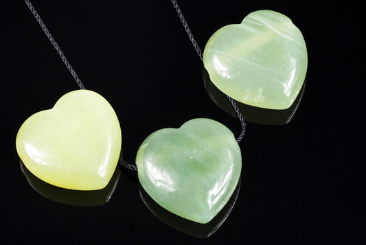 Three polished heart-shaped jade pendants in pale yellow and green tones hang from dark cords on a black reflective surface.
