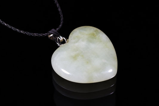 A heart-shaped jade pendant with pale green and white tones hangs from a dark cord against a black background.
