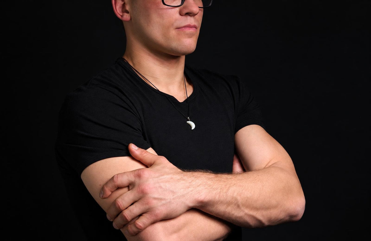 A man in a black t-shirt wears a necklace with a white, crescent-shaped pendant against a dark background.
