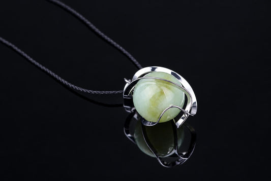 A silver-toned wire pendant holds a smooth, pale green sphere on a dark cord against a black reflective surface.