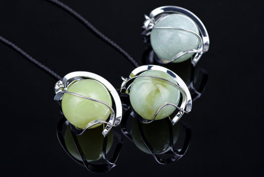 Three silver-toned pendants with pale green spheres on black cords, arranged on a dark reflective surface.