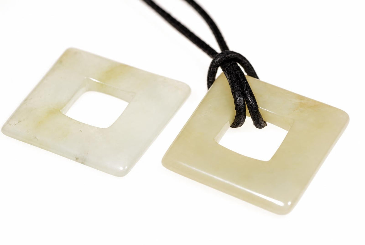 Two square jade pendants with central holes; one has a black cord, both on a white surface.