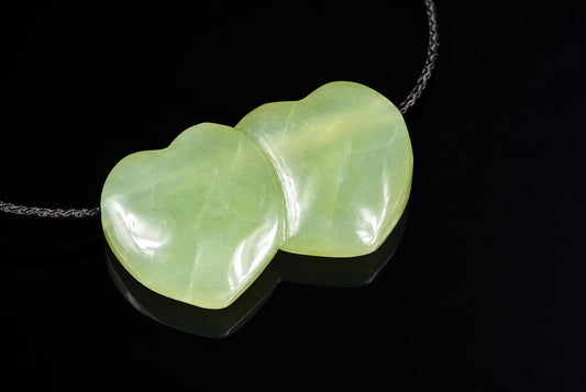 A pale green, heart-shaped pendant with a smooth surface hangs on a dark cord against a black background.